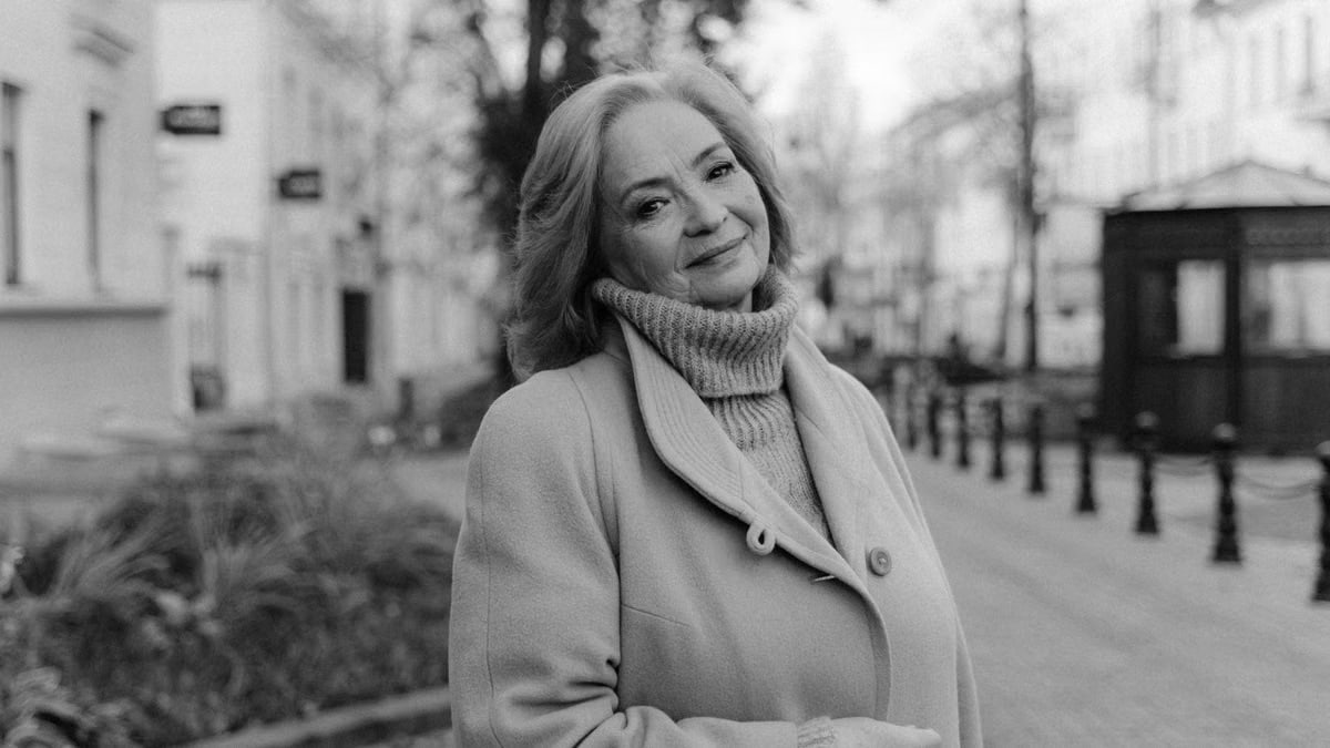 A stylish senior woman standing on a city sidewalk, smiling warmly in grayscale photography.